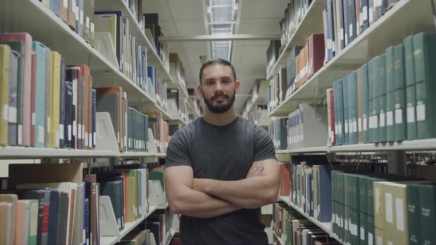 A man stands with his arms crossed in the aisle of a library with shelves full of books