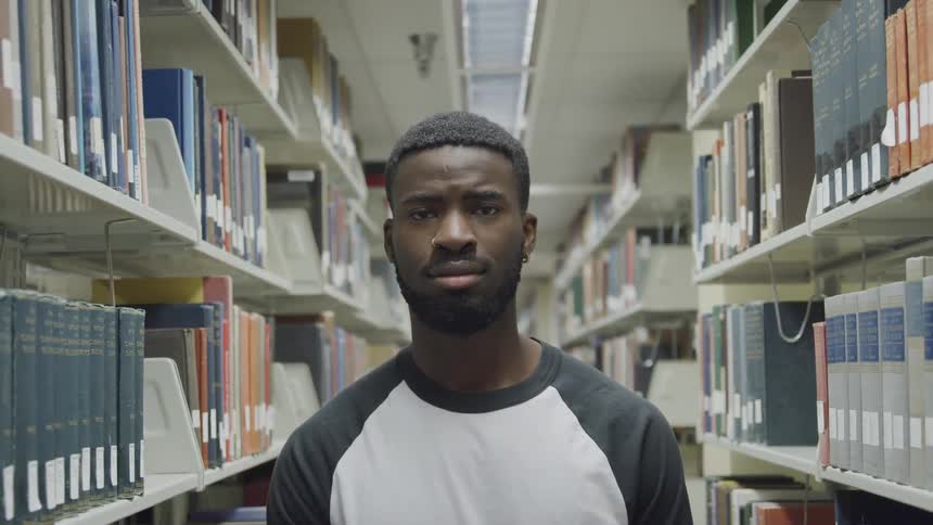 Black man with a white and black sweatshirt faces the camera with eye contact in the library