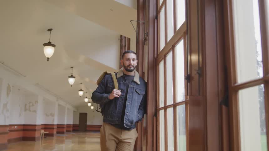 Young man wearing a jacket carrying a backpack stands in the hallway of a university