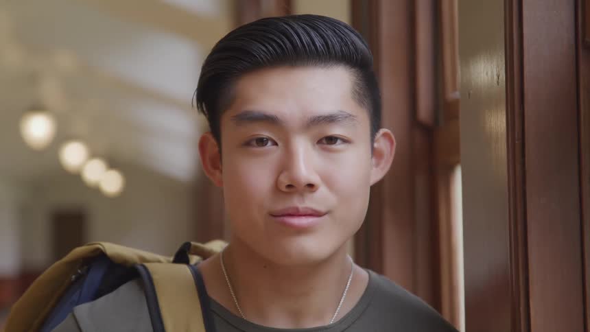 A young man poses for the camera while holding a backpack and standing near a window