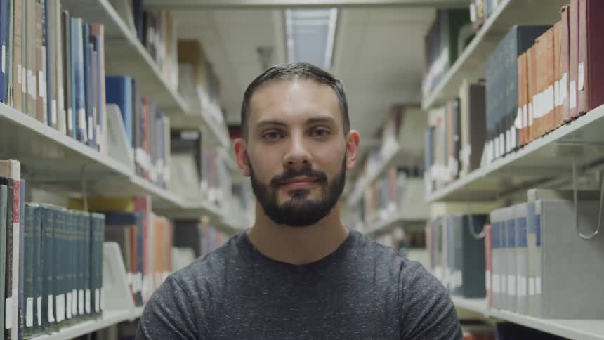 A man with dark hair and a beard and mustache is standing between shelves loaded with books in a library