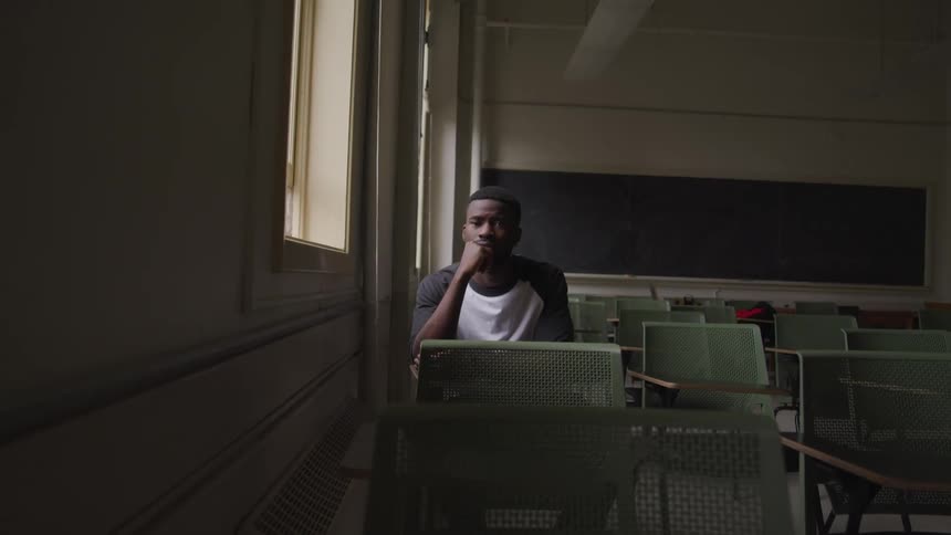 A young male sits at a school desk in a dark classroom