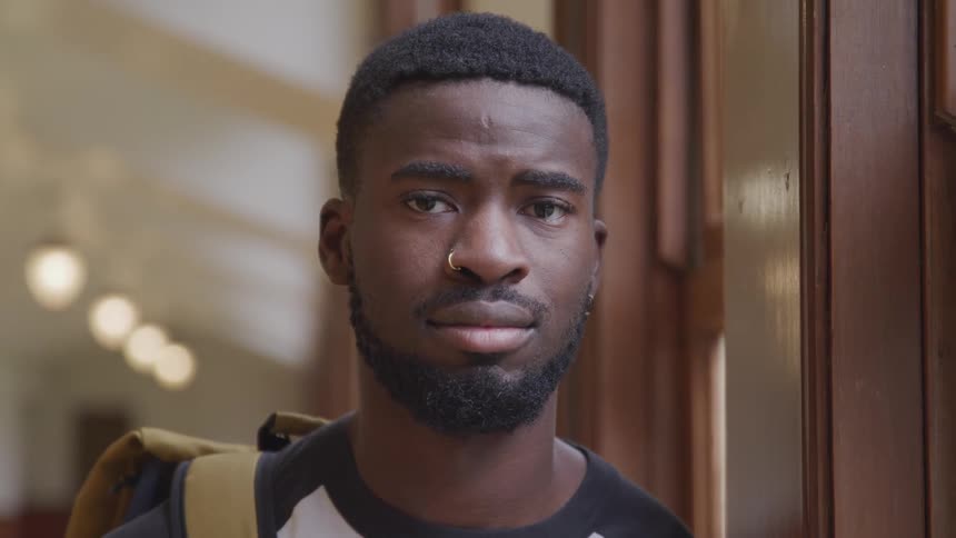 Student carrying a backpack next to a window on a hallway looks at the camera