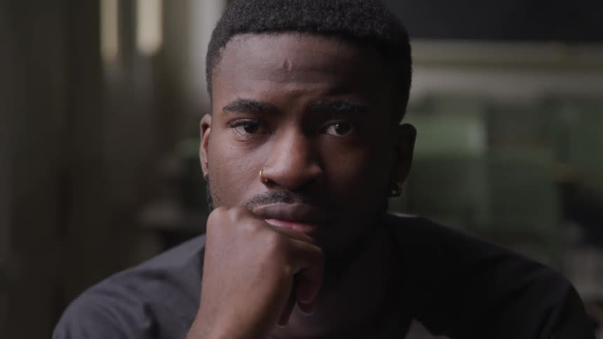 A young black man inside a building rests his chin in his hand