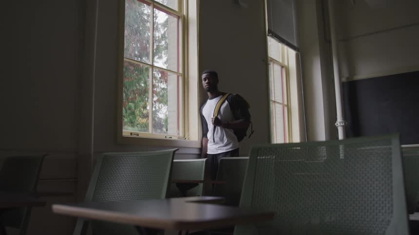 A college student stands next to a window in an empty classroom
