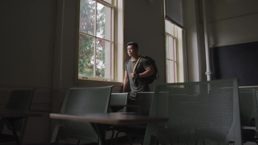 A teenager in a classroom leans against the wall and looks out the window