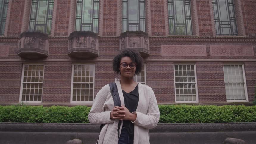 Black woman with short curly hair glasses and a backpack looks at the camera smilingly in front of a building