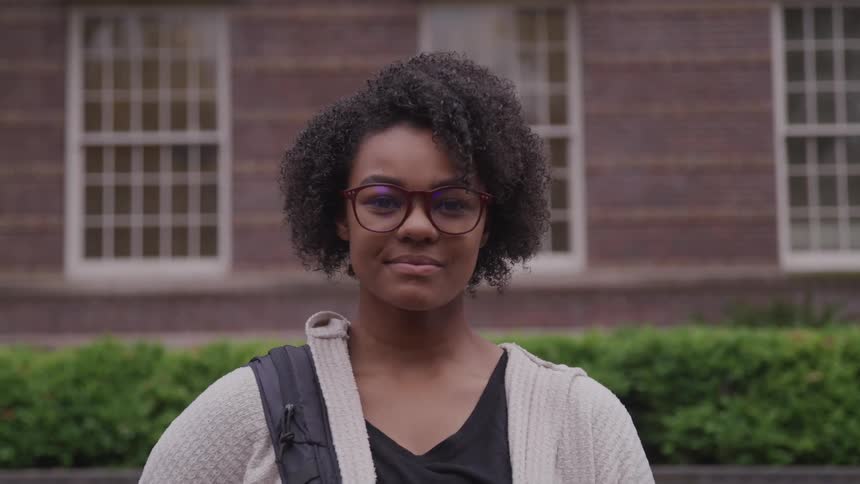 Black woman with a short curly hair glasses and a backpack faces the camera smilingly with eye contact in front of a building