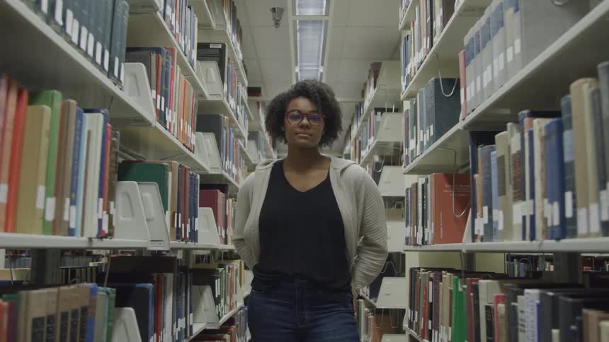 A young black woman stands in the aisle between bookshelves in a library