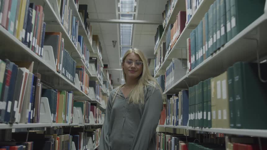 A woman with a sweater and glasses standing and smiling between bookshelves