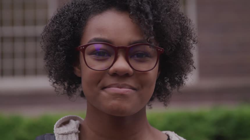 Black woman with short curly hair and glasses looks at the camera smilingly in front of a building