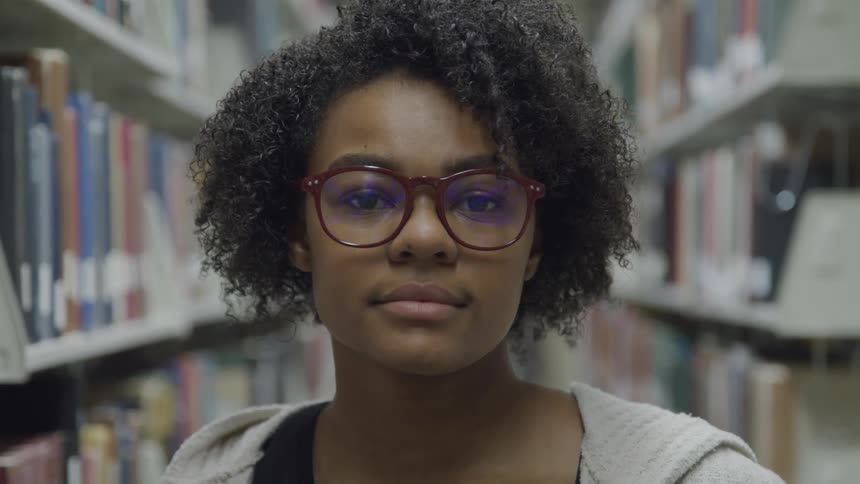 Black woman with curly hair and glasses faces the camera with eye contact in the library between bookshelves