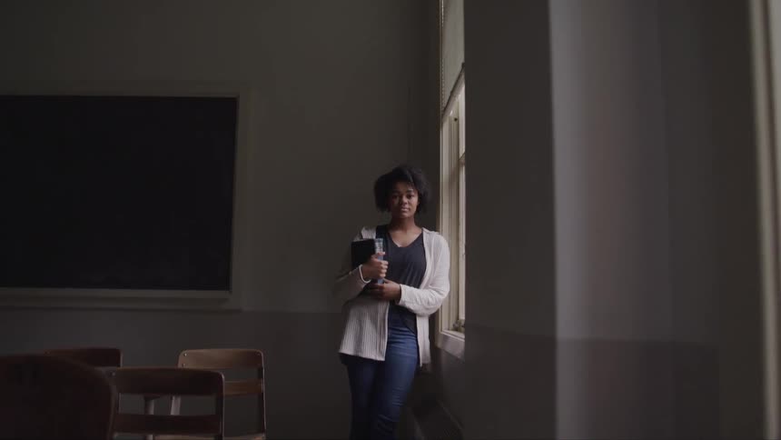 A young woman holds a binder as she poses next to a window in an empty classroom