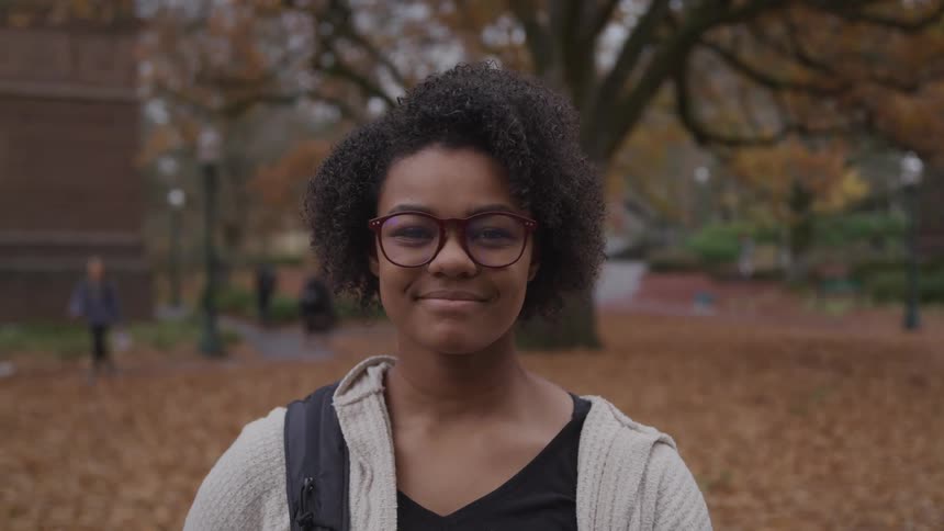 A young black woman wearing a backpack as she stands among fallen leaves shifts her gaze to look into the camera