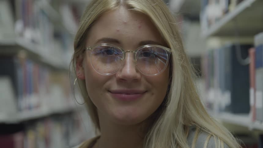 A young woman with long blonde hair with glasses stands within a library