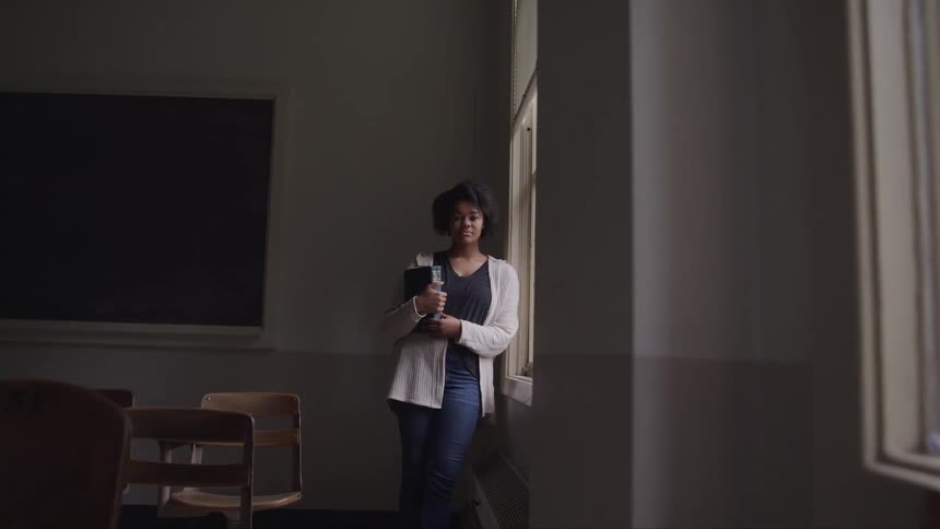 Black woman with curly hair holds a book and stands next the windows in the dim lit classroom