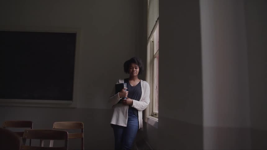 Black woman with curly hair holds a book and looks at the camera in dim lit classroom