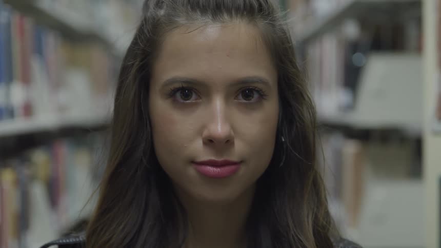 Young woman standing on a library looks at the camera and starts smiling
