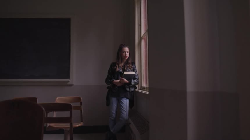 Woman with a leather jacket and jeans stands next to a window and looks at the camera in a dim lit classroom