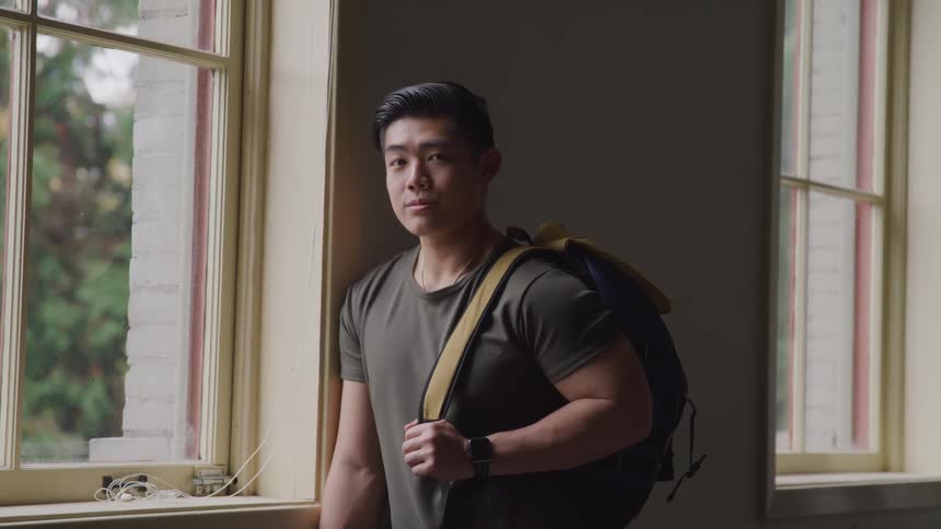 Muscular asian man leaning against a wall looks out the window and turns his head to face the camera in a classroom