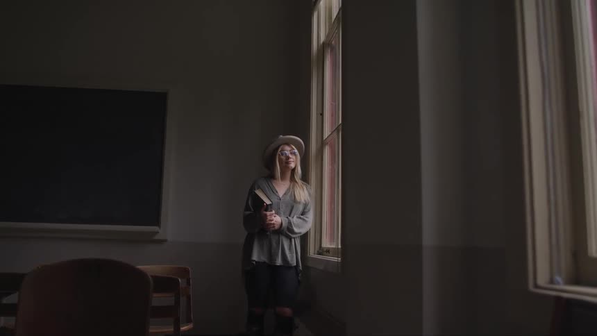 A white girl with blonde hair wearing a hat is standing next to a window in a classroom holding a book
