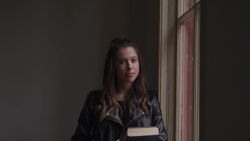 A teenage girl holding a textbook stands in a classroom and looks out the window