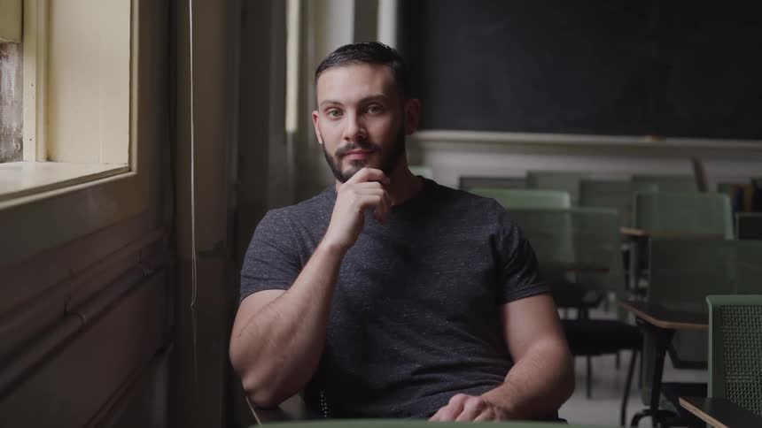Man with a beard and a black t shirt sits in a chair and looks at the camera in classroom