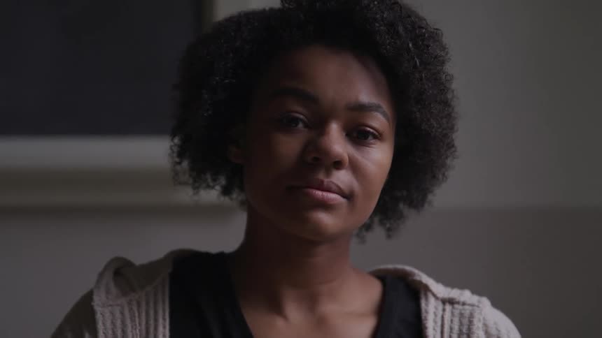Black girl with curly hair faces the camera with eye contact in the dim lit classroom