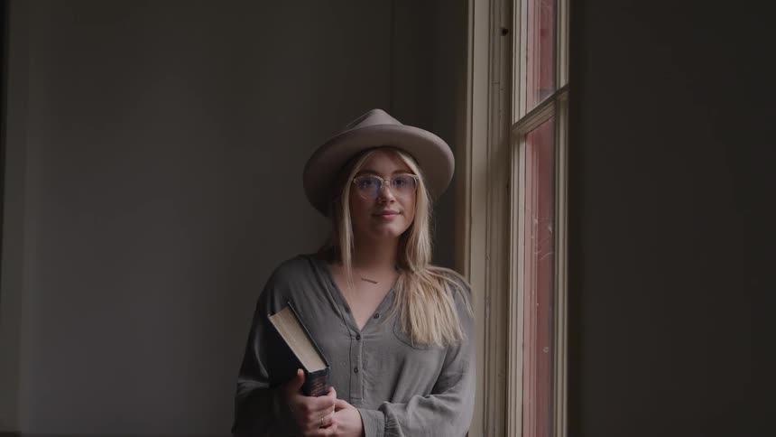 A blonde caucasian girl with a tan hat on stands next to a window in a dimly lit room with a book in her hand