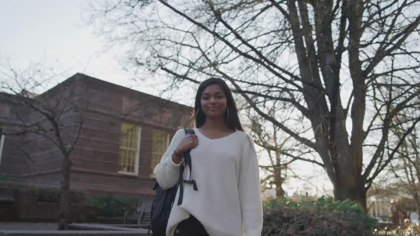 Dark skinned girl with straight long hair and a white sweatshirt stands and looks at the camera smilingly at sunset