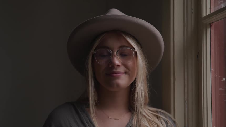 A woman wearing a large felt hat and glasses poses indoors next to a window