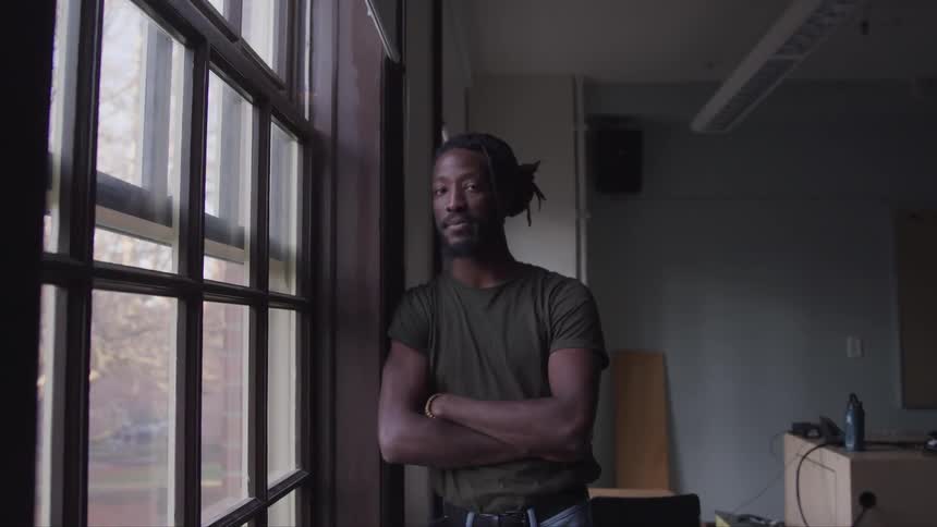 Black man with a beard and a green t shirt stands next to the window and looks at the camera