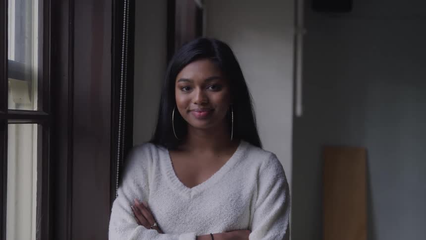 A young black woman stands indoors next to a large window while smiling