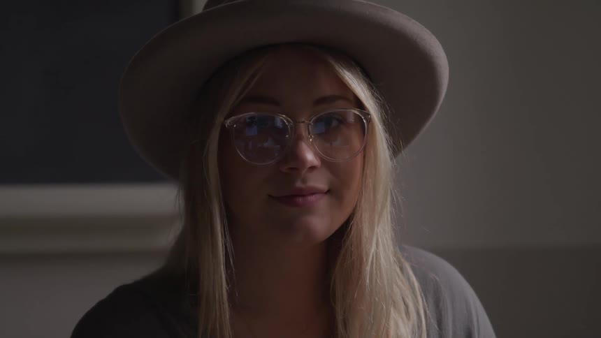 Close up of a blonde woman wearing glasses and a hat in a room looking at the camera