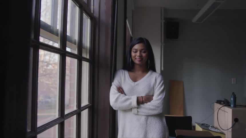 Young woman standing next to a window in a room looks at the camera smiling