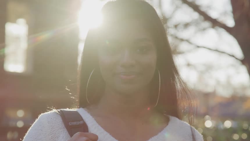 Dark skinned girl with hoop earrings and a white sweater smiles and looks at the camera as the sun shines brightly in the background
