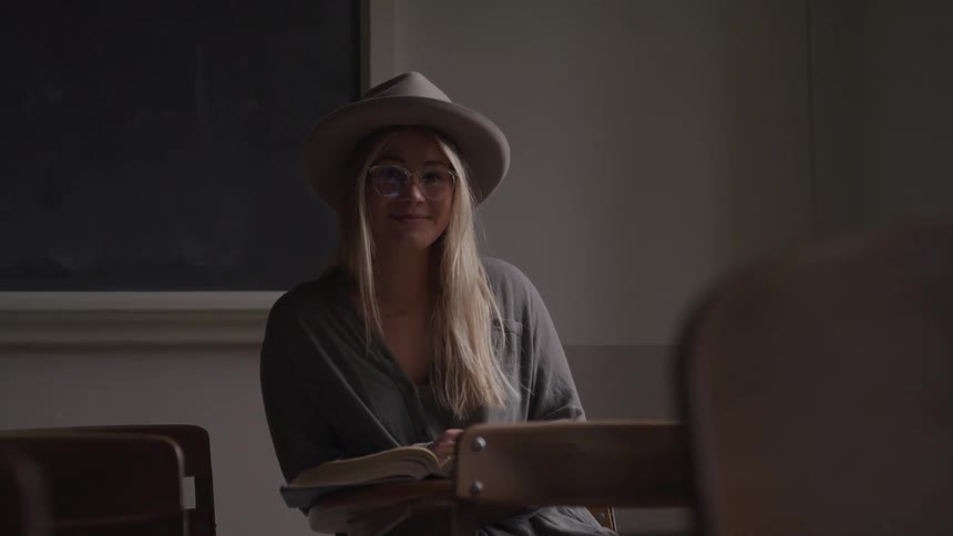 A young woman sitting in a classroom looks up from reading a book