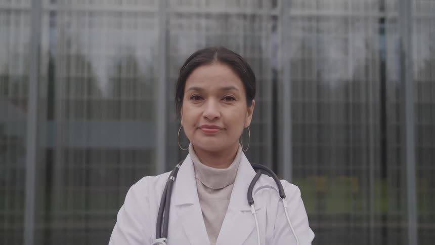 A woman with a lab coat and a stethoscope standing and staring with a window in the background