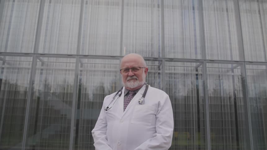 A elderly doctor stands outside of a building during the day and looks into the camera