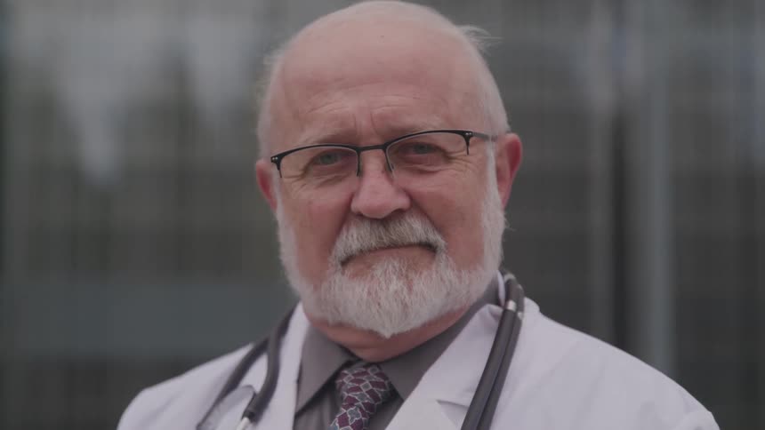 An elderly white doctor with balding white hair is standing in front of a building with his stethoscope draped around his neck over his white lab coat