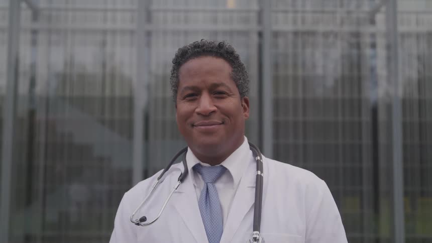 A doctor poses outside a hospital with a stethoscope around his neck