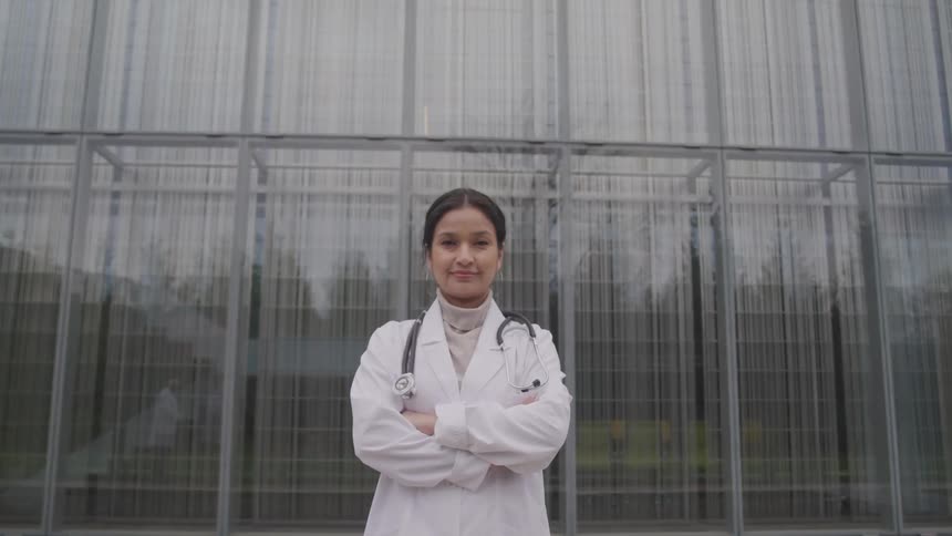 A doctor in a white coat poses with her arms crossed in front of large glass windows at a hospital