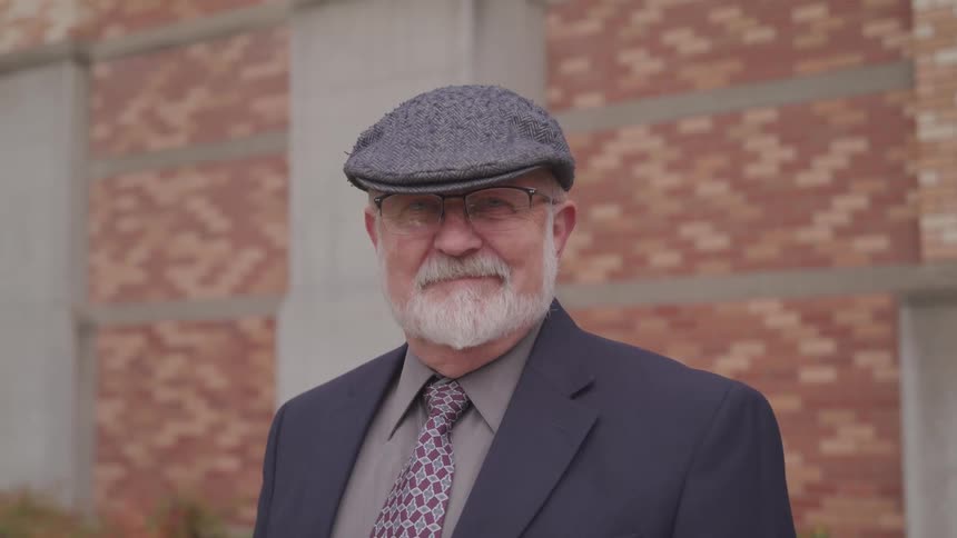 Elderly man wearing a suit and a hat looks at the camera in front of a building