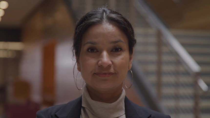 Woman with a blazer and hoop earrings faces the camera with eye contact in a large room