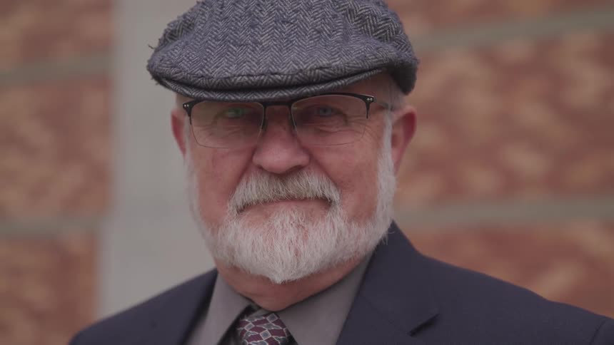 Elderly man with white beard wearing hat and suit looks into camera