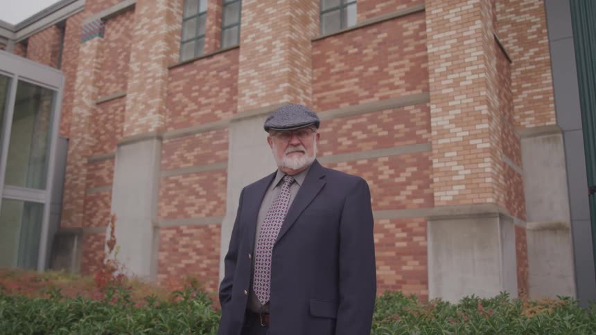 Elderly man with a beard a grey flat cap and a suit stands and looks at the camera in front of a building