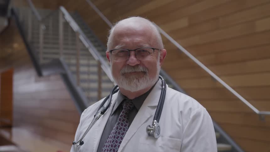 An elderly doctor with white hair stands looking ahead with a white lab coat and stethoscope