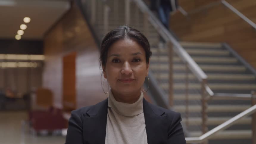 An indian woman in formal clothing smiles into the camera while standing inside a building
