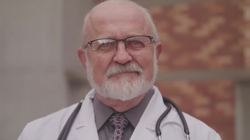 A grinning elderly doctor turns his head to look into the camera while standing outdoors during the day
