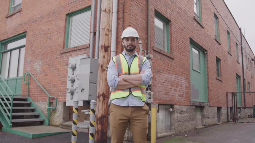 A man wearing a construction vest and hard hat stands outside of a brick building